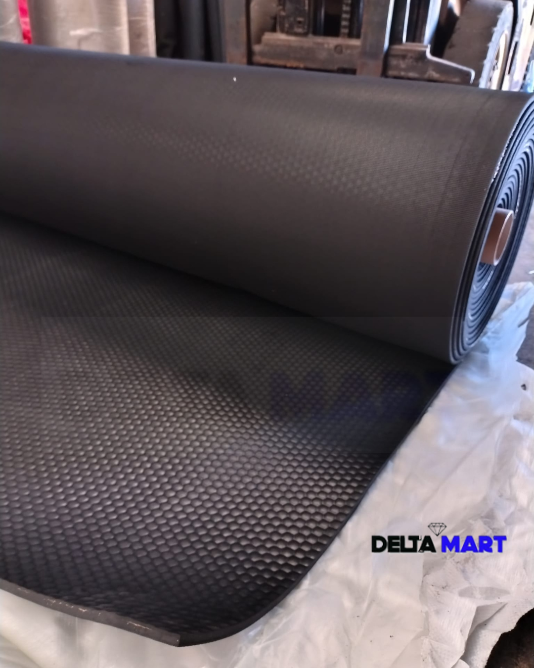 Large roll of black textured conveyor belt or mat partially unrolled on a white sheet, factory setting in the background, Delta Mart watermark.