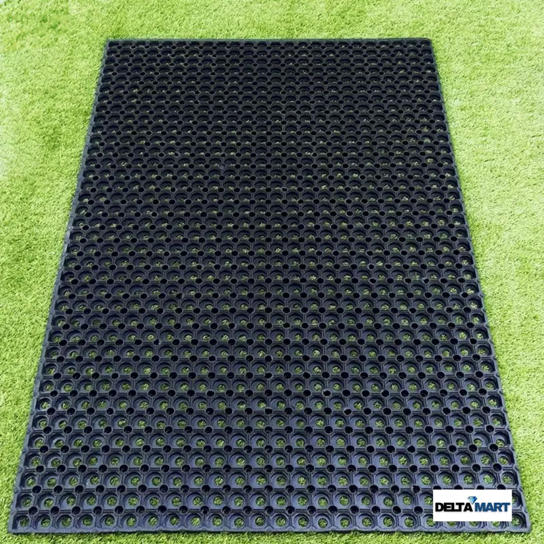 Black perforated rubber mat with circular holes laid on green artificial turf; DeltaMart logo in the corner.