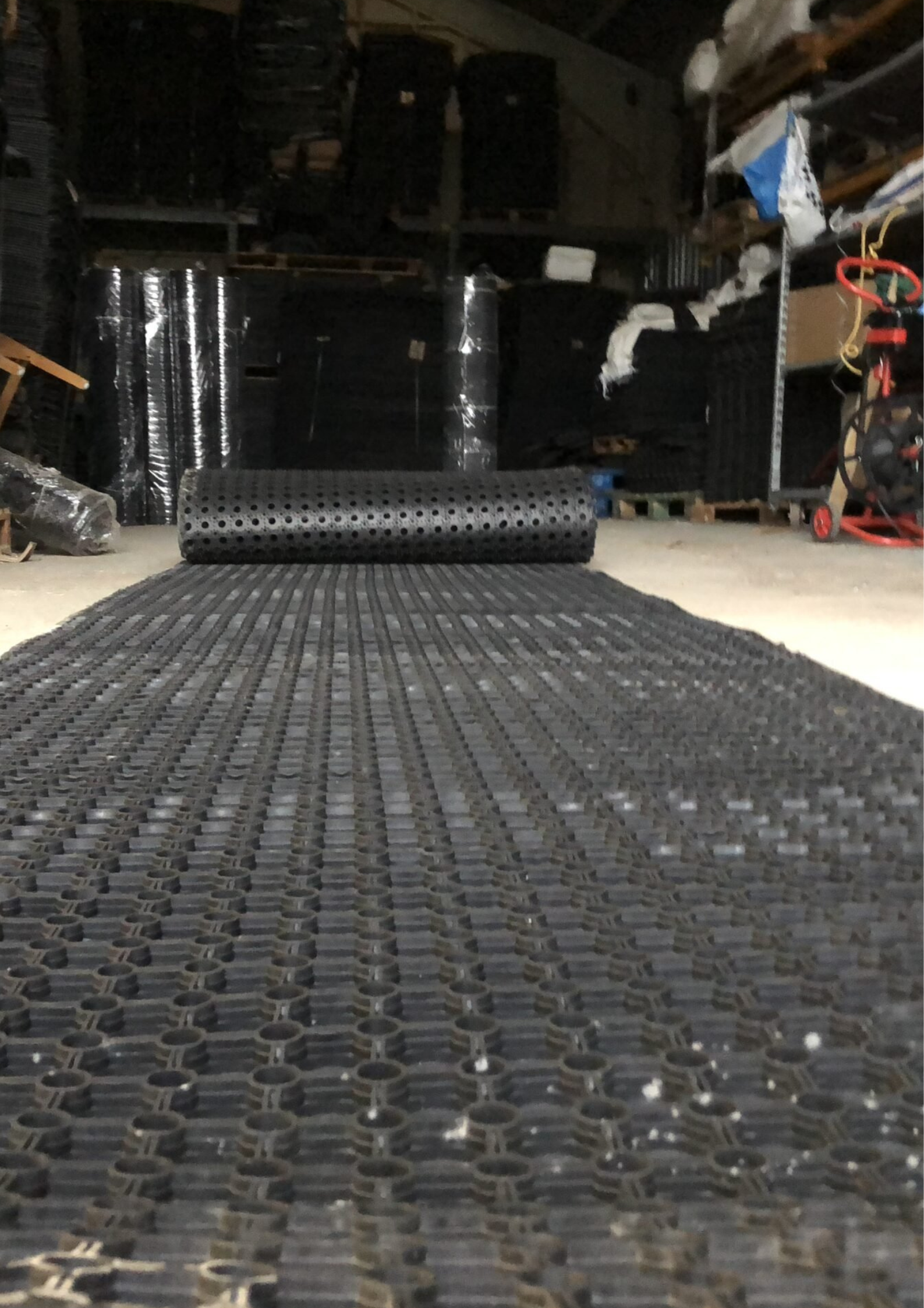 Close-up of black perforated interlocking anti-fatigue mats rolled up on a workshop floor, with tools and clutter in the background.