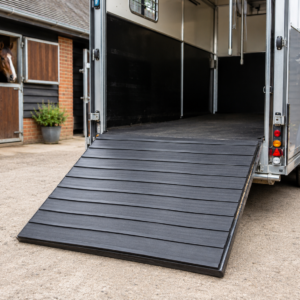 Open horse trailer with a lowered ramp, parked near a stable; a horse peers out of a stall window nearby.