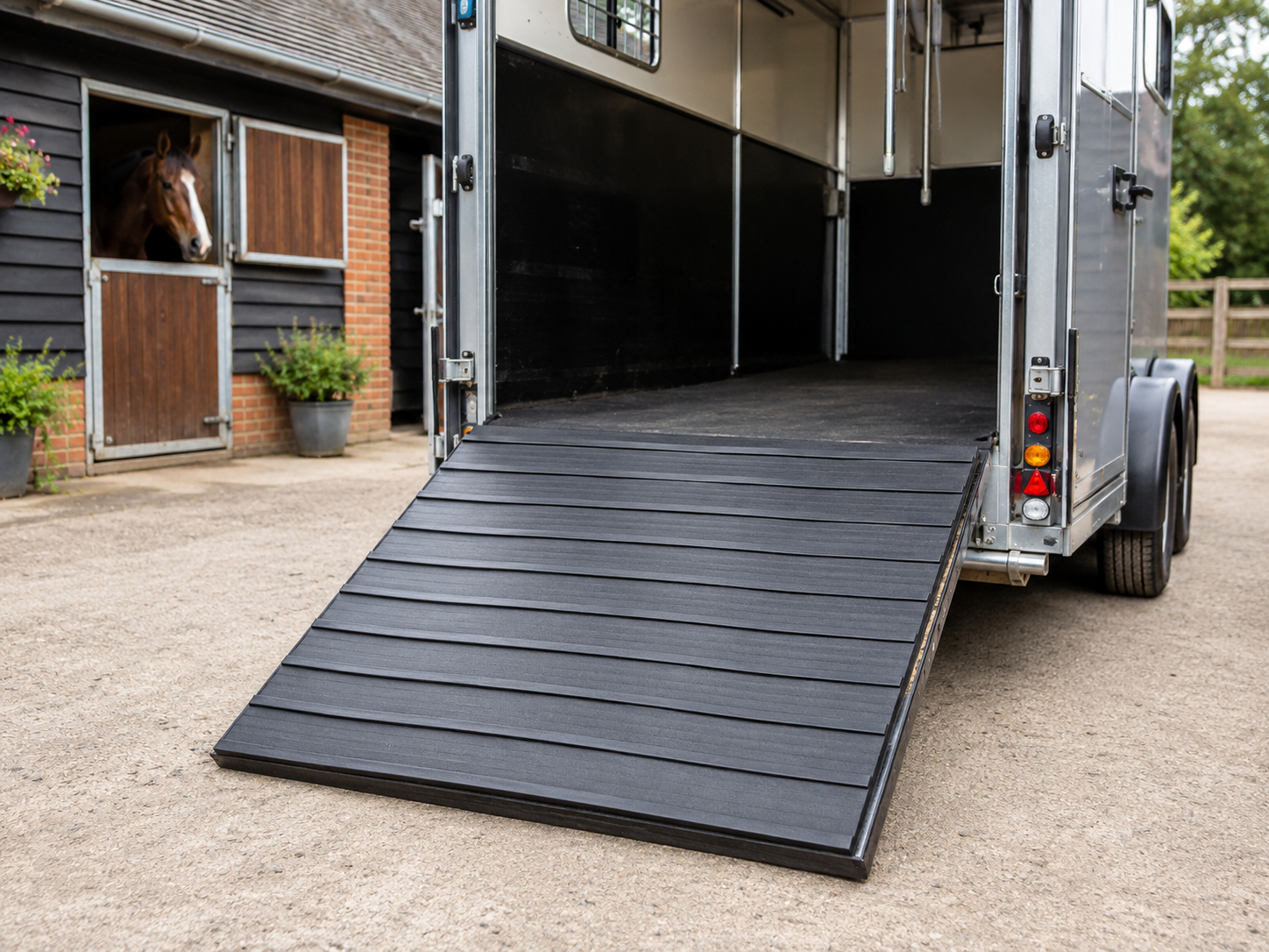 Open horse trailer with a lowered ramp, parked near a stable; a horse peers out of a stall window nearby.