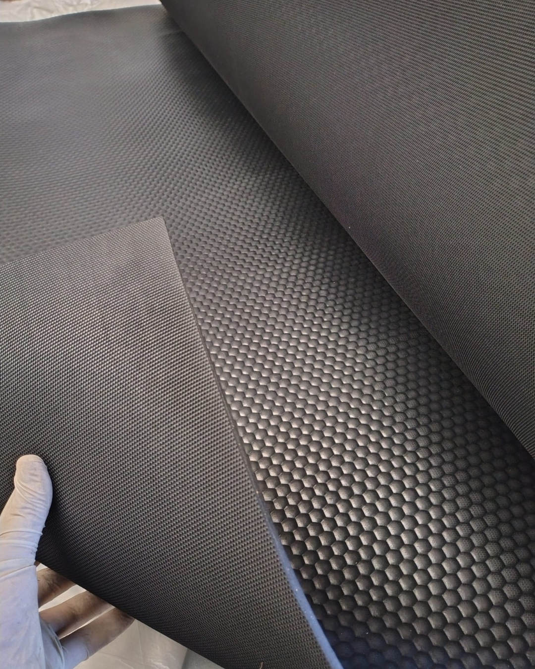 Hands in a glove lift a sheet of black textured rubber with a honeycomb pattern.