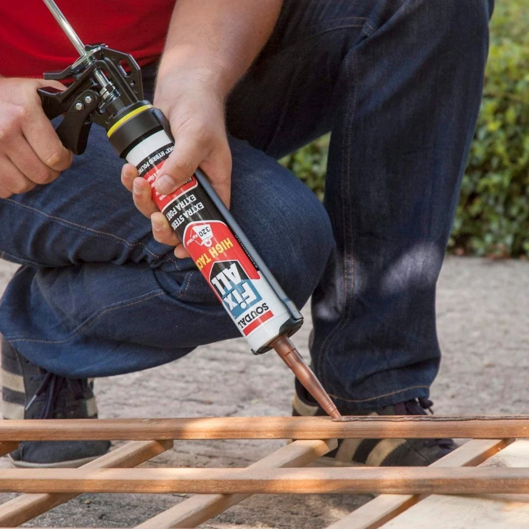 Person in jeans kneeling, applying caulk from a tube onto a wooden frame.